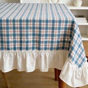 Puede incluir: Un mantel a cuadros azules y blancos con líneas rojas. El mantel tiene un ribete blanco con volantes en el borde inferior. El texto "BROKEN FLOWER GIFTS" está impreso en el mantel.