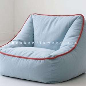 Pode incluir: Uma cadeira de saco azul claro com acabamento vermelho. A cadeira tem formato arredondado e é feita de tecido macio. O texto "Brokenflower Gifts" é visível na cadeira.