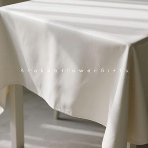 May include: A cream-coloured tablecloth on a table, with soft folds and shadows. The fabric has a subtle texture. The text "Brokenflower Gifts" is printed on the tablecloth.