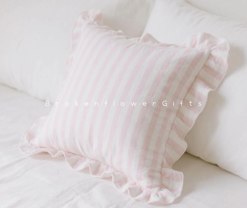 May include: A square pink and white striped pillow with ruffled edges. The pillow is on a white bed with other white pillows. The text "Brokenflower Gifts" is printed on the pillow.