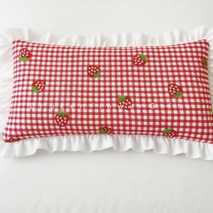 May include: A rectangular cushion featuring a red and white gingham design, decorated with strawberry motifs. The cushion is trimmed with a white frill, offering a decorative touch for a bed or sofa.