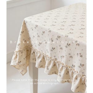 May include: Cream-colored tablecloth with a delicate floral pattern and ruffled edges. The fabric is adorned with small, scattered flowers in shades of brown and gray. The text "Brokenflower Gifts" is visible on the tablecloth.