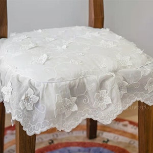 May include: White lace chair cushion with a ruffled edge and floral embroidery. The cushion is on a wooden chair with a brown finish.