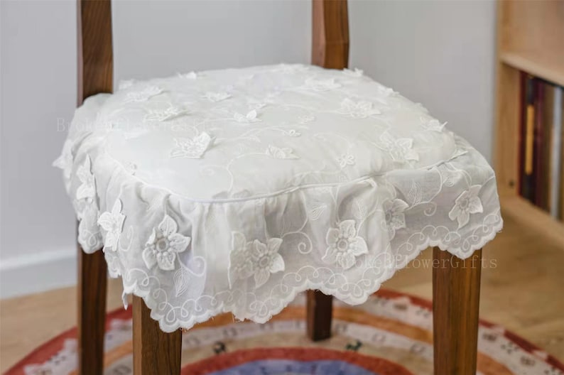 May include: White lace chair cushion with a ruffled edge and floral embroidery. The cushion is on a wooden chair with a brown finish.