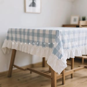 Op de afbeelding: Een lichtblauw en wit geruit tafelkleed met een witte gefranjerde rand. Het tafelkleed is over een houten tafel gedrapeerd. De tekst "BrokenFlower Gifts" is zichtbaar op het tafelkleed.