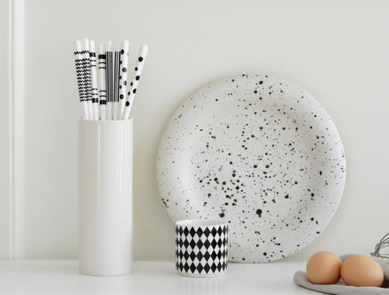 May include: A white ceramic vase with a bunch of black and white patterned straws inside. A white ceramic plate with black speckles, a white ceramic mug with a black and white checkered pattern, and two brown eggs are on a white surface.