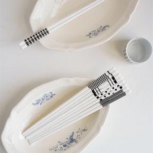 May include: A set of white chopsticks with black and white geometric patterns. The chopsticks are arranged in a bowl and on a plate with a blue and white floral design.