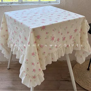 Puede incluir: Un mantel cuadrado color crema con un estampado floral de rosas rosas. El mantel tiene un borde con volantes y está sobre una pequeña mesa blanca. El texto "Brokenflower Gifts" está impreso en el mantel.