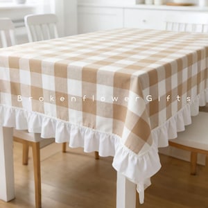 Puede incluir: Un mantel a cuadros beige y blanco con un borde con volantes blancos. El mantel está sobre una mesa blanca, con sillas blancas en el fondo. El texto "Brokenflower Gifts" está impreso en el mantel.