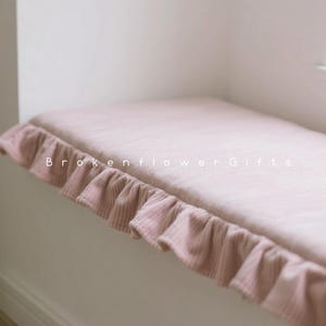 May include: A pale pink window seat cushion with a frilled edge. The cushion is made of a corduroy-like fabric and is placed on a white window ledge. The text "Brokenflower Gifts" is printed on the cushion.