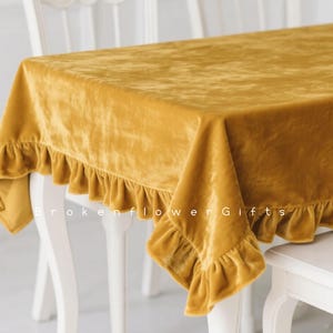 Puede incluir: Mantel de terciopelo amarillo mostaza con bordes con volantes. El mantel está sobre una mesa y una silla blancas. El texto "Brokenflower Gifts" es visible en el mantel.