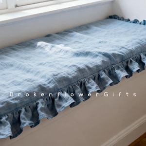 May include: A pale blue bench cushion with a frilled border. The cushion is made from a soft, textured material and sits on a white bench. The text "Broken Flower Gifts" is printed on the bottom of the image.