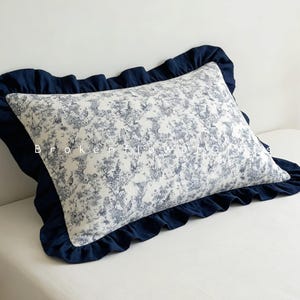 May include: A rectangular pillow with a white and blue floral pattern, framed by a navy blue ruffled border. The pillow is placed on a white surface. The text "Broken Flowers" is visible on the pillow.