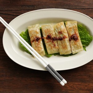 May include: Four golden brown dumplings served on a white oval plate with a bed of green lettuce. A pair of white chopsticks with black accents are placed on the plate.