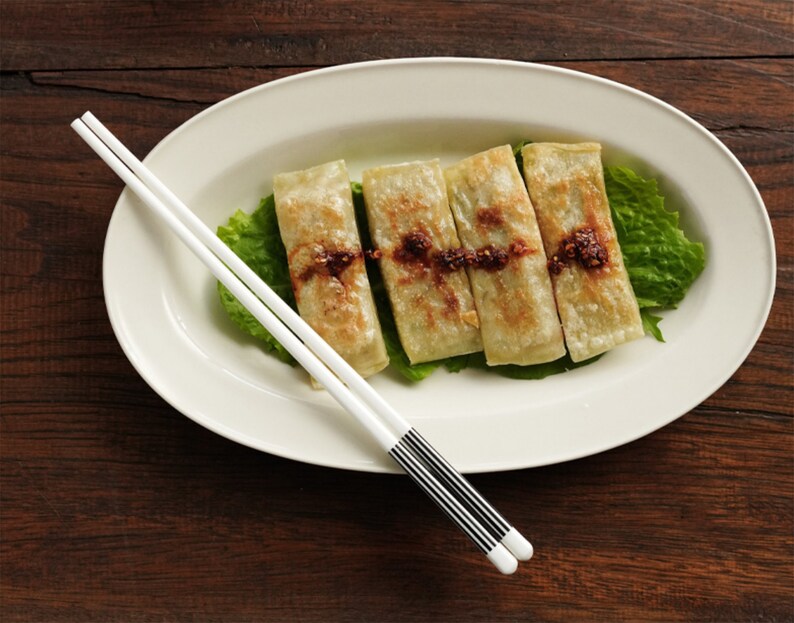 May include: Four golden brown dumplings served on a white oval plate with a bed of green lettuce. A pair of white chopsticks with black accents are placed on the plate.
