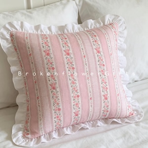 Könnte beinhalten: Ein quadratisches Zierkissen mit einem rosa-weiß gestreiften Muster, kleinen Blumenmotiven und einem weißen Rüschenrand. Der Text BrokenflowerGifts ist in einer einfachen weißen Schrift mittig auf dem Kissen aufgedruckt.