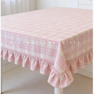 Può includere: Una tovaglia a quadri rosa e bianca con bordo a balze. La tovaglia è su un tavolo bianco. Il testo "Brokenflower Gifts" è stampato sulla tovaglia.