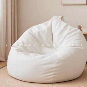 May include: A large, white beanbag chair. The chair is made of a soft, smooth fabric and is filled with small beads. The beanbag is on a beige rug. The text "Brokenflower Gifts" is visible on the beanbag.