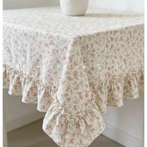 Puede incluir: Un mantel cuadrado con un estampado floral de flores rosas y verdes sobre un fondo crema. El mantel tiene un borde con volantes y el texto "Brokenflower Gifts" es visible.