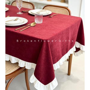 May include: A burgundy rectangular tablecloth with white ruffled trim. The tablecloth is set on a wooden table with gold-colored cutlery, white plates, and clear glassware. The text "Brokenflower Gifts" is visible.