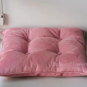 May include: A pink velvet floor cushion with a tufted design. The cushion has a square shape and is filled with soft padding.