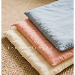 Pode incluir: Três almofadas quadradas e macias em cores pastel: azul claro, pêssego e creme. As almofadas estão empilhadas umas sobre as outras. O texto "Brokenflower Gifts" é visível na parte inferior. As almofadas são feitas de tecido texturizado.