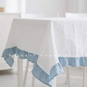 May include: White tablecloth with a light blue frilled edge. The tablecloth is draped over a table with white legs. The text "Brokenflower Gifts" is printed on the tablecloth.