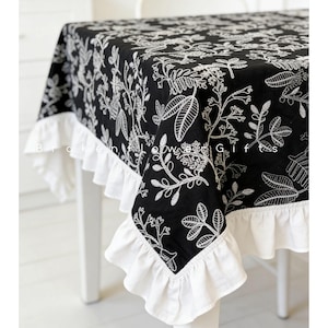 Könnte beinhalten: Schwarze Tischdecke mit weißer Blumenstickerei und weißem Rüschensaum. Die Tischdecke ist über einem weißen Tisch drapiert. Der Text "Broken Bloom Gifts" ist auf der Tischdecke zu sehen.