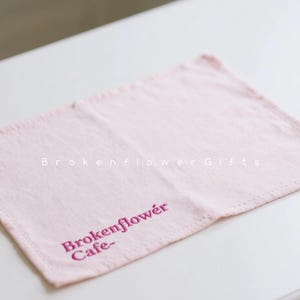 Könnte beinhalten: Ein hellrosa rechteckiges Tuch mit gesticktem Text. Der Text lautet "Brokenflower Gifts" und "Brokenflower Cafe" in dunklerem Rosa. Das Tuch liegt auf einer weißen Oberfläche.