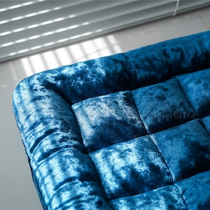 Può includere: Un cuscino blu in velluto capitonné con un motivo quadrato. Il cuscino è realizzato in un tessuto morbido e soffice e ha una sensazione lussuosa.