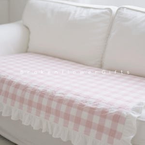 Può includere: Un divano bianco con una coperta a quadri rosa e bianca. La coperta ha un bordo a balze e il testo "Brokenflower Gifts" è stampato sopra.