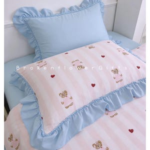 May include: A light blue and pink bedding set with a teddy bear pattern. The set includes a pillow with a ruffled edge and the text "Brokenflower Gifts". The pillow has a pink and white striped pattern with teddy bears and red hearts.