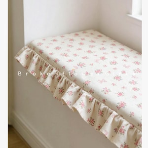 Könnte beinhalten: Ein Fensterbankkissen mit einem Blumenmuster aus rosa Rosen und Schmetterlingen auf cremefarbenem Hintergrund. Das Kissen hat einen gerüschten Rand aus passendem Blumenstoff. Der Text "Brokenflower" ist auf dem Kissen zu sehen.