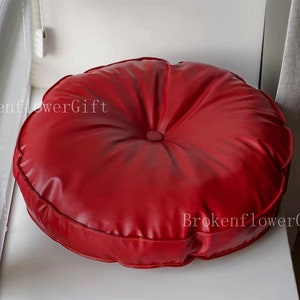 May include: A round, red, leather cushion with a button in the center. The cushion is sitting on a white surface.