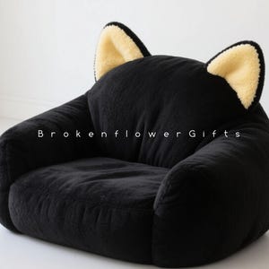 May include: A black plush cat-shaped chair with two upright ears featuring a cream-colored interior. The chair has a rounded seat and armrests. The text "Brokenflower Gifts" is printed on the chair.