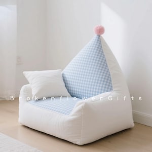 Puede incluir: Una silla infantil blanca de textura suave con respaldo y asiento de cuadros azules y blancos. Una pequeña almohada blanca descansa sobre el brazo. Un pompón rosa adorna la parte superior. El texto "Brokenflower Gifts" es visible.