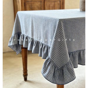 Peut inclure: Une nappe à carreaux bleus et blancs avec un bord à volants. La nappe est drapée sur une table en bois. Le texte "Broken Flower Gifts" est imprimé sur la nappe.
