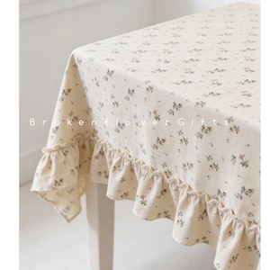 May include: Cream-colored tablecloth with a delicate floral pattern and ruffled edges. The fabric features small, scattered flowers in shades of brown and gray. The text "Brokenflower Gifts" is printed on the tablecloth.