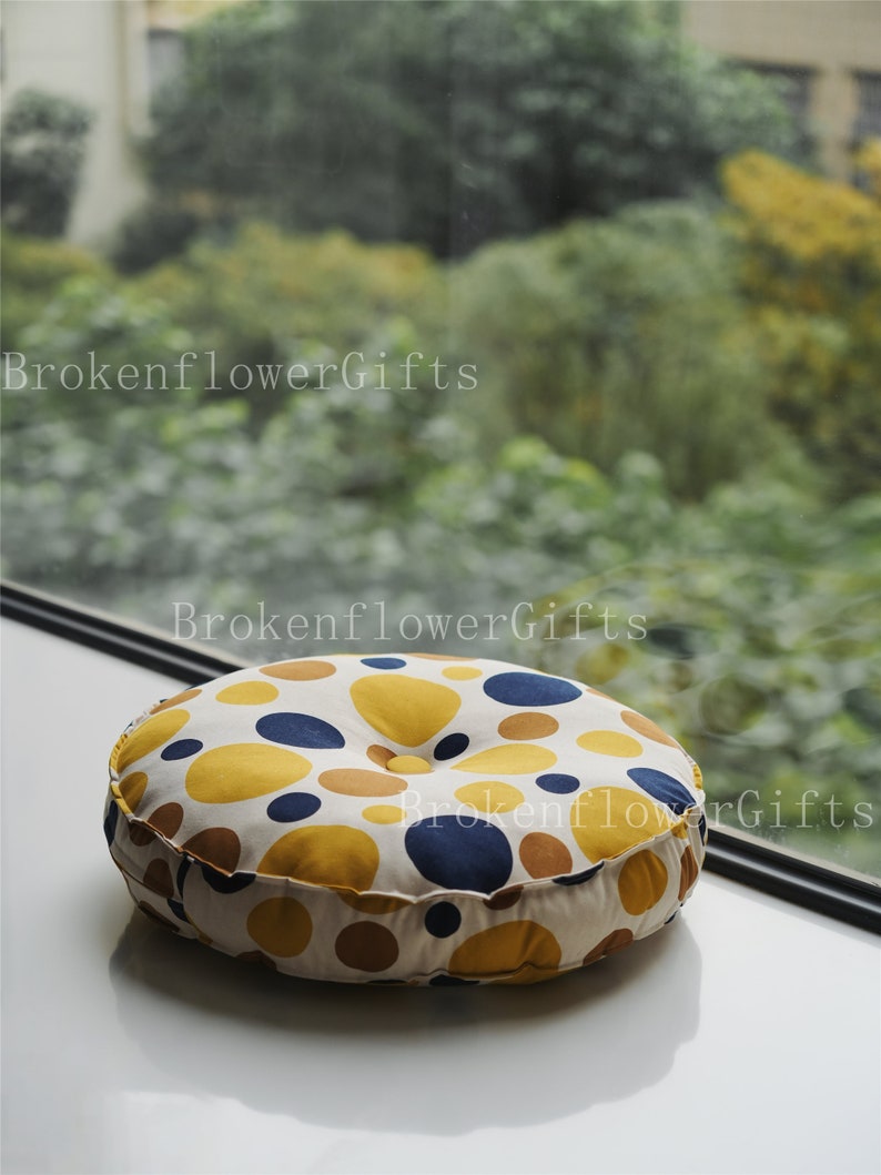 Op de afbeelding: Een rond, cr&egrave;mekleurig kussen met een knoop in het midden. Het kussen is bedekt met een stof met een stippenpatroon in geel, bruin en blauw.