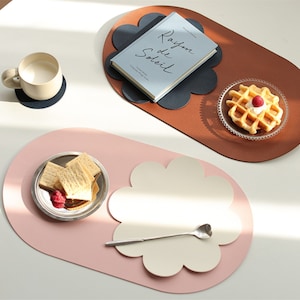 Op de afbeelding: Twee ovale leren placemats met een bloemvormig ontwerp in het midden. De placemat links is roze met een witte bloem, en de placemat rechts is bruin met een zwarte bloem. De placemat rechts heeft een boek met de titel "Rayon de Soleil" erop. De placemat links heeft een bord met crackers en frambozen erop. De placemat rechts heeft een bord met wafels met een framboos erop.