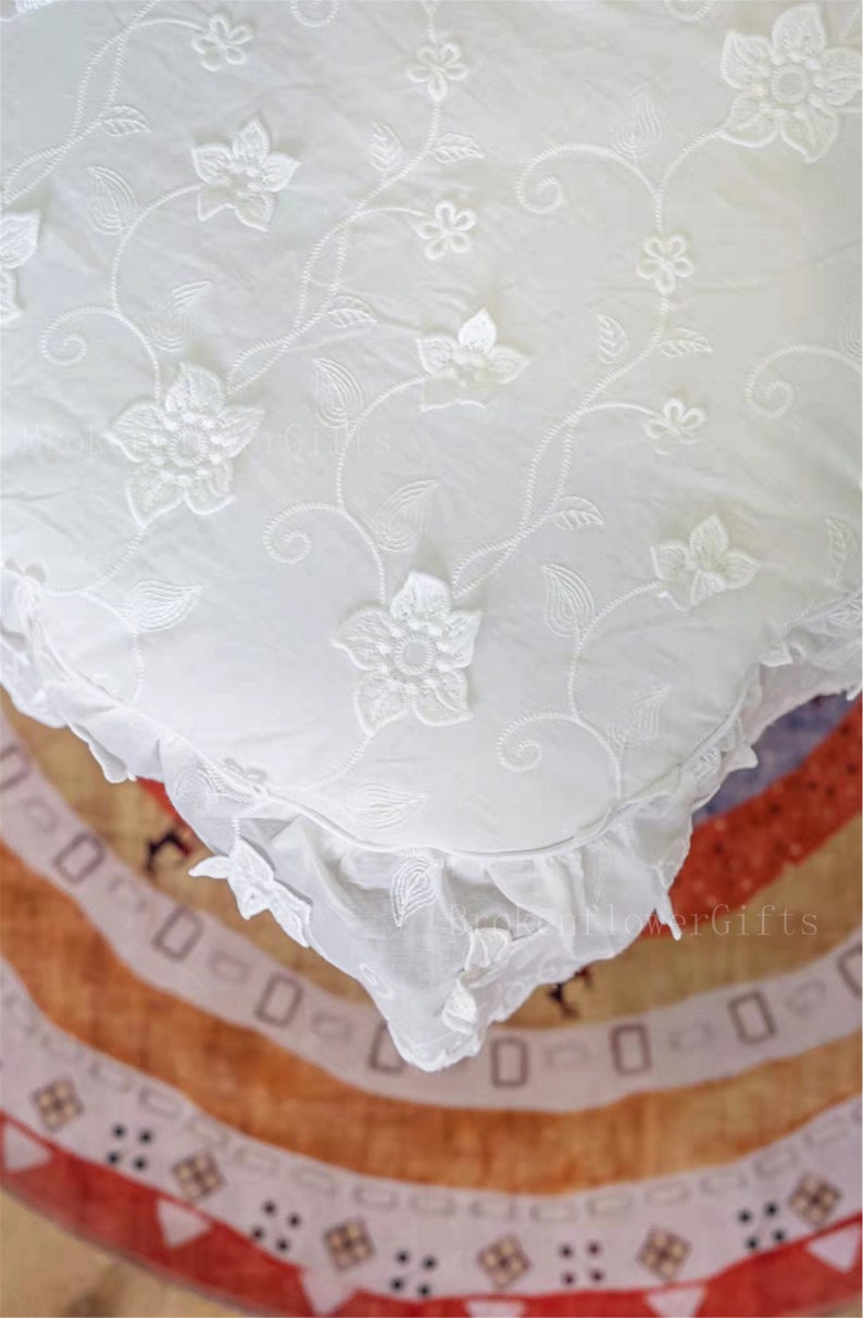 May include: White cotton pillow cover featuring a delicate floral embroidery design. The pillow cover has a ruffled edge and is made from a soft, lightweight fabric.