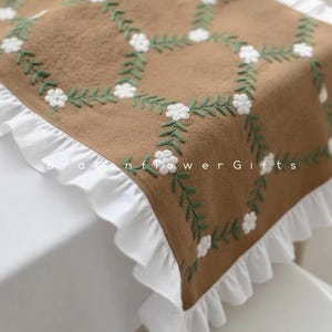 Puede incluir: Una tela marrón con flores blancas bordadas y hojas verdes dispuestas en un patrón de cuadrícula. La tela tiene un borde blanco con volantes. El texto "Broken flower Gifts" es visible en la tela.