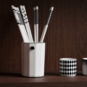 May include: A white ceramic octagonal vase with a black knob on the side, holding a set of black and white patterned chopsticks. Two white ceramic cups with black geometric patterns are on the shelf below.