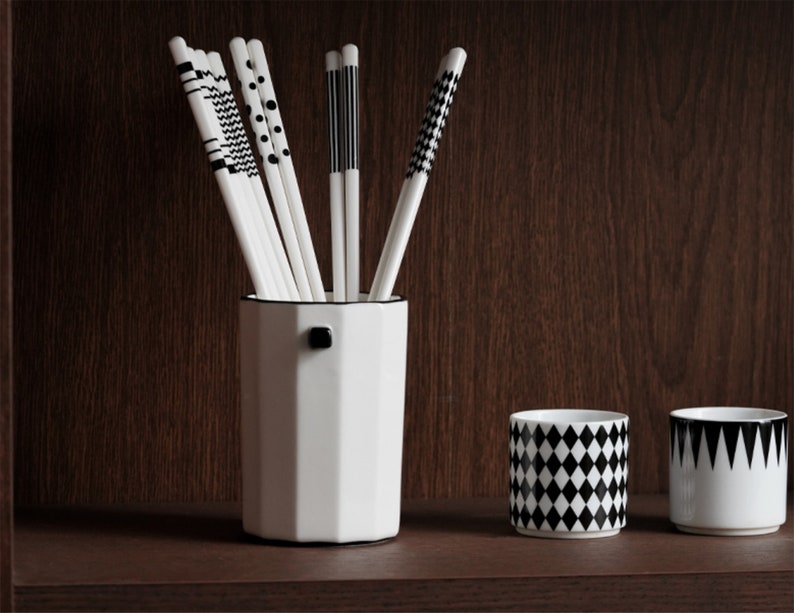 May include: A white ceramic octagonal vase with a black knob on the side, holding a set of black and white patterned chopsticks. Two white ceramic cups with black geometric patterns are on the shelf below.