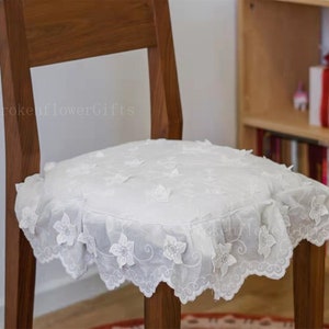 May include: A white chair cushion with a delicate lace design and floral embroidery. The cushion is placed on a wooden chair.