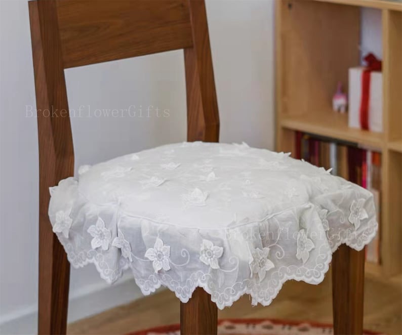 May include: A white chair cushion with a delicate lace design and floral embroidery. The cushion is placed on a wooden chair.