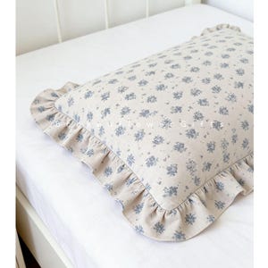May include: A rectangular cushion with a floral pattern in light blue and beige. The cushion has a frilled edge and the text "B. Flower Gifts" is visible. The cushion is on a white bed.