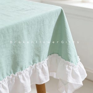 Pode incluir: Uma toalha de mesa verde claro com uma borda branca com babados. A toalha de mesa está drapeada sobre uma mesa. O texto "Brokenflower Gifts" está impresso no tecido.