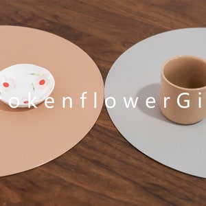 May include: Two round placemats, one light brown and one light gray, with a small white plate with a pink floral design on the brown placemat and a light brown ceramic mug on the gray placemat. The text "BrokenflowerGift" is printed in white on the placemats.