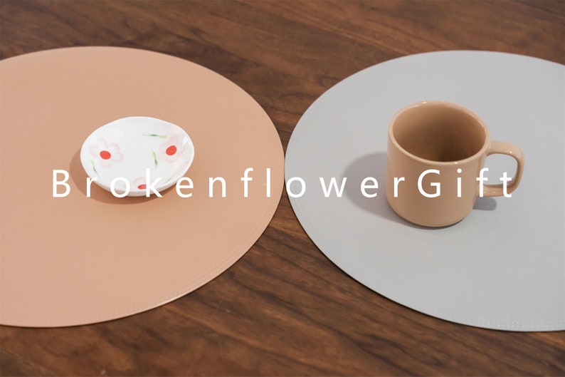 May include: Two round placemats, one light brown and one light gray, with a small white plate with a pink floral design on the brown placemat and a light brown ceramic mug on the gray placemat. The text "BrokenflowerGift" is printed in white on the placemats.
